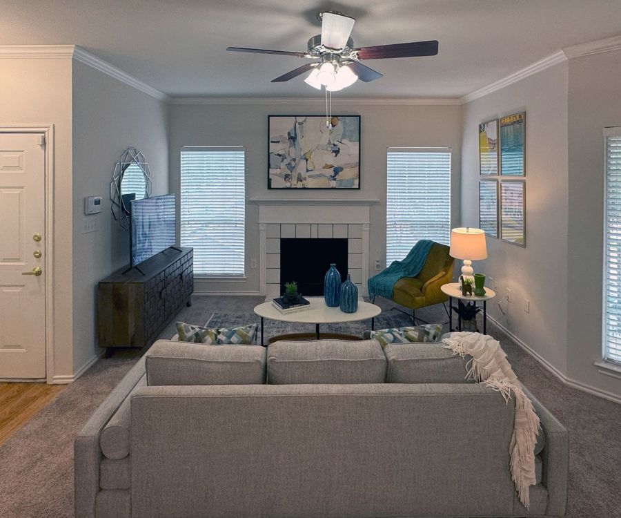 Cozy living room with a gray sofa, TV, ceiling fan, and artwork above a fireplace. Lamp and decor enhance the ambiance.