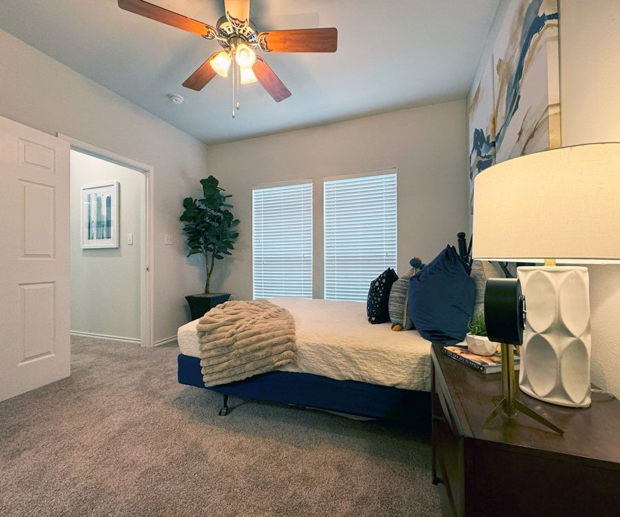 Cozy bedroom with a bed, decorative pillows, a lamp on a nightstand, ceiling fan, and artwork on the walls.
